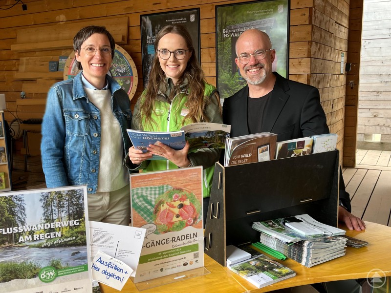 Ursula Schuster, Andrea Kralik und Robert Kürzinger am Messestand der Ferienregion und Nationalparkverwaltung Bayerischer Wald auf der Landesgartenschau in Furth im Wald. Drei Personen stehen lächelnd an einem Informationsstand aus Holz. Auf dem Tisch vor ihnen befinden sich zahlreiche Broschüren und Flyer zur Ferienregion Nationalpark Bayerischer Wald sowie Plakate zur Landesgartenschau und touristischen Angeboten.