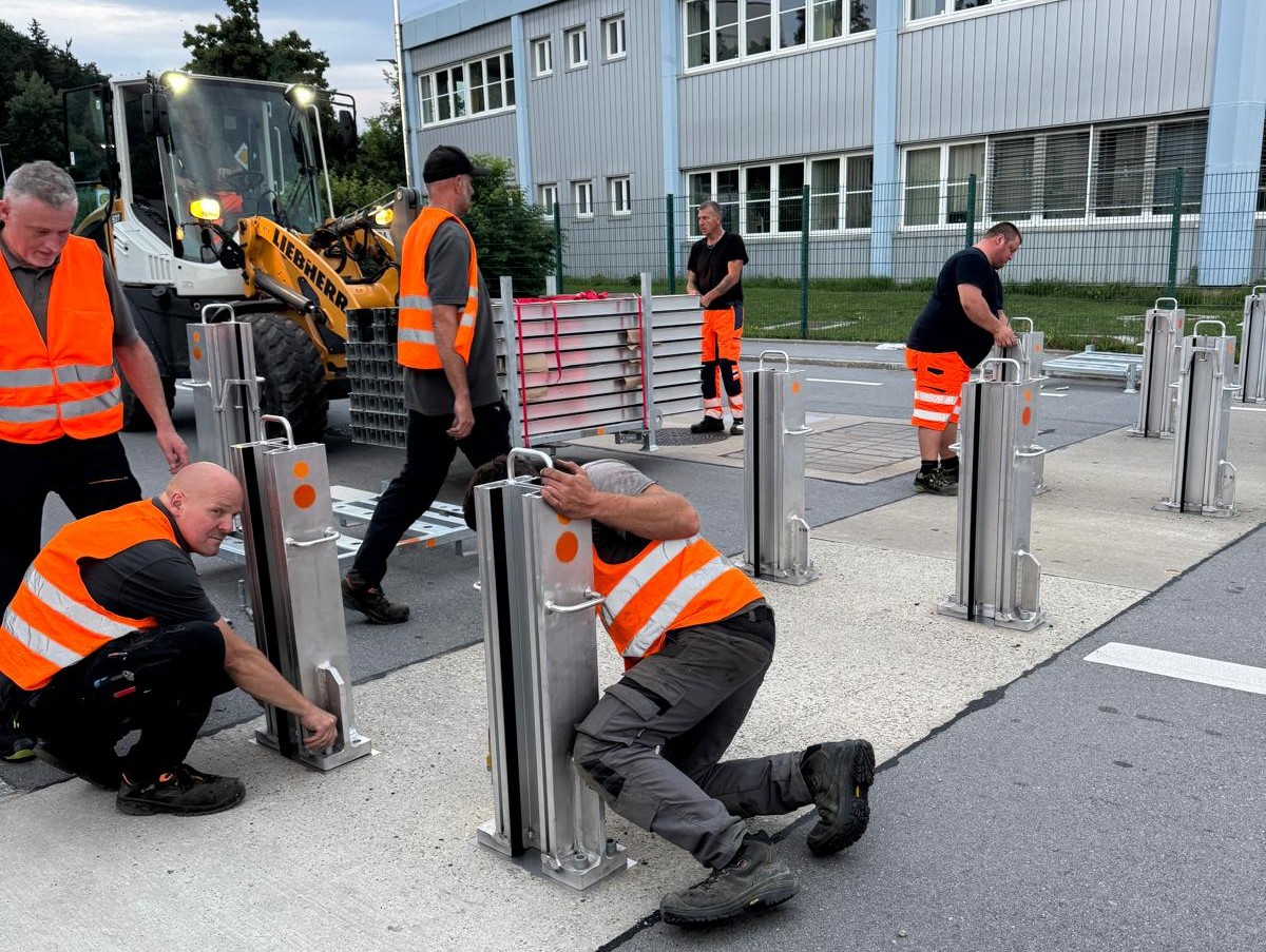 Zweiter Probeaufbau des Hochwasserschutzes Lindau/Sulzsteg Mehrere Arbeiter in orangefarbenen Warnwesten montieren in den frühen Morgenstunden mobile Hochwasserschutzelemente auf einer Straße vor einem Industriegebäude. Im Hintergrund ist ein Bagger im Einsatz.