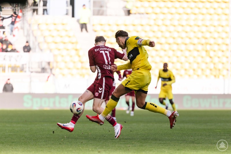 Zweikampfstark: Der Jahn Regensburg zeigte sich beim 2:0-Auswärtssieg in Aachen kompromisslos und kampfstark. Ein Spieler im weinroten Trikot und ein Spieler in Gelb kämpfen direkt am Ball. Beide setzen sich mit vollem Körpereinsatz ein, im Hintergrund erkennt man die gelbe Tribüne und weitere Spieler.