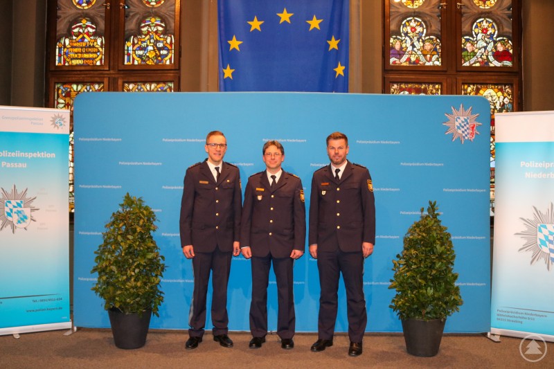 Amtswechsel bei der Grenzpolizeiinspektion Passau: Der neue Leiter Polizeioberrat Sebastian Feucht (rechts) übernimmt die Führung von Polizeidirektor Thomas Ritzer (Mitte), im Beisein von Polizeipräsident Roland Kerscher (links). Drei Polizeibeamte in Uniform stehen für ein offizielles Foto vor einer blauen Pressewand mit dem Logo des Polizeipräsidiums Niederbayern. Im Hintergrund sind eine Europaflagge und bunte Glasfenster zu sehen. Rechts und links im Bild stehen Banner der Grenzpolizeiinspektion Passau sowie zwei grüne Pflanzen.