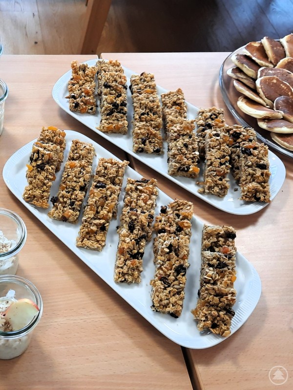 Mehrere l&auml;ngliche M&uuml;sliriegel aus Haferflocken, N&uuml;ssen und Trockenfr&uuml;chten liegen auf wei&szlig;en Tellern auf einem Tisch, daneben sind kleine Pfannkuchen und Gl&auml;ser mit Joghurt zu sehen.