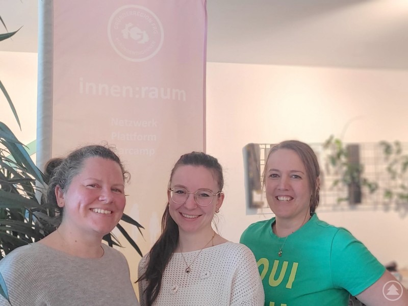 Stephanie Fichtl l. und Jasmin Binder r. vom GreG FRG werden bei der Organisation auch von Viktoria Kohlpaintner m. von der Initiative gemeinwohlorientiert.com unterstützt. Drei Frauen stehen lächelnd vor einem Banner des innenraum Netzwerks und posieren für ein Gruppenfoto.
