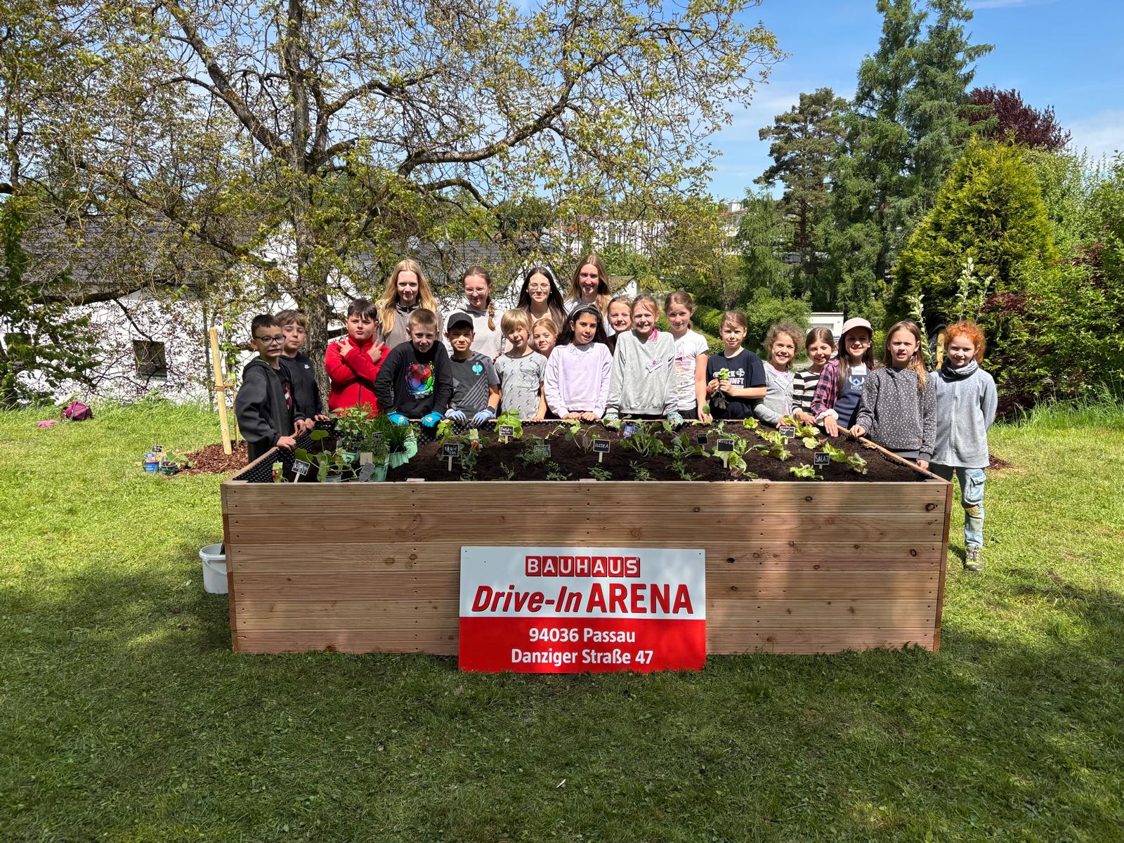 Gemeinsam aktiv für Nachhaltigkeit: Schülerinnen und Schüler präsentieren ihr fertig bepflanztes Beet im Garten des Seniorenheims Mariahilf. Eine Gruppe von Schülerinnen und Schülern steht vor einem frisch bepflanzten Hochbeet im Garten des Seniorenheims Mariahilf. Ein Bauhaus-Schild weist auf den Sponsor hin.