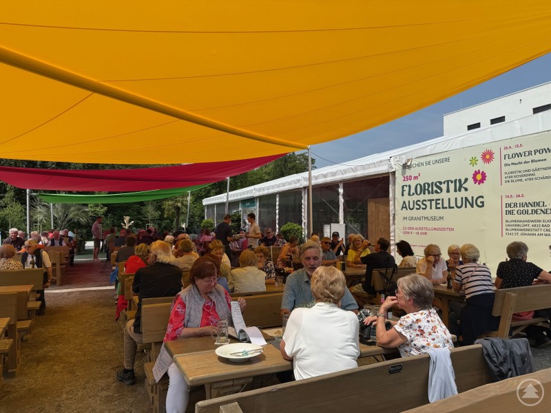 Gemütliches Beisammensein beim Mittagessen im Gastgarten des Bio-Wirtes auf dem Gartenschaugelände. Zahlreiche Besucherinnen und Besucher sitzen im Schatten bunter Sonnensegel an langen Holzbänken und genießen ein gemeinsames Mittagessen.