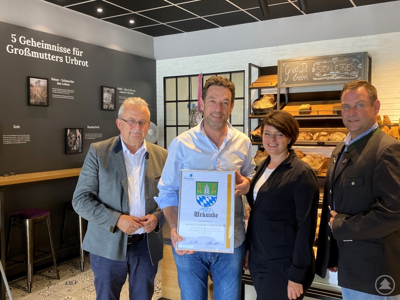 Landrat Josef Laumer (links) überreicht gemeinsam mit Wirtschaftsförderer Martin Köck (rechts) eine Urkunde an Markus und Stefanie Steinleitner zum 65-jährigen Betriebsjubiläum der Bäckerei Steinleitner. Vier Personen stehen in einer Bäckerei vor einem Regal mit Broten. Zwei Männer und eine Frau überreichen einem weiteren Mann eine Urkunde. Im Hintergrund ist eine Wand mit dem Text „5 Geheimnisse für Großmutters Urbrot“ zu sehen.