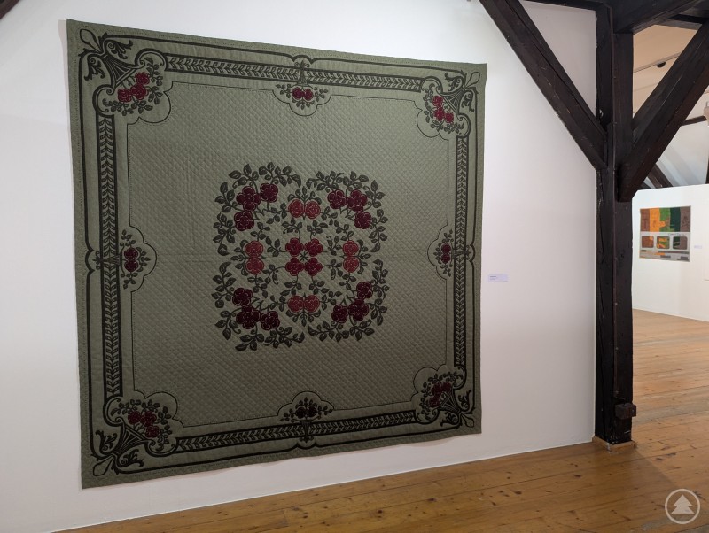 Traditionell gestalteter Quilt mit floralem Muster in der Ausstellung „Tradition bis Moderne“ im Stadtmuseum Deggendorf. Ein großer, quadratischer Quilt in Grün- und Rottönen mit floralen Mustern hängt an einer weißen Wand in einem Museumsraum mit Holzboden.