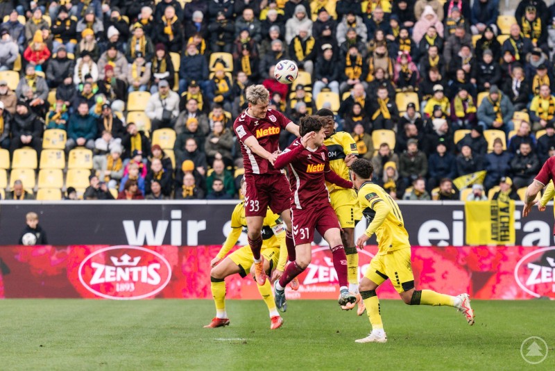Zwei Regensburger Spieler springen im Strafraum hoch und versuchen, den Ball per Kopf zu klären oder zum Tor zu bringen. Zahlreiche Spieler beider Mannschaften kämpfen um den Ball, das Publikum ist in gelb-schwarz auf den Rängen zu sehen.