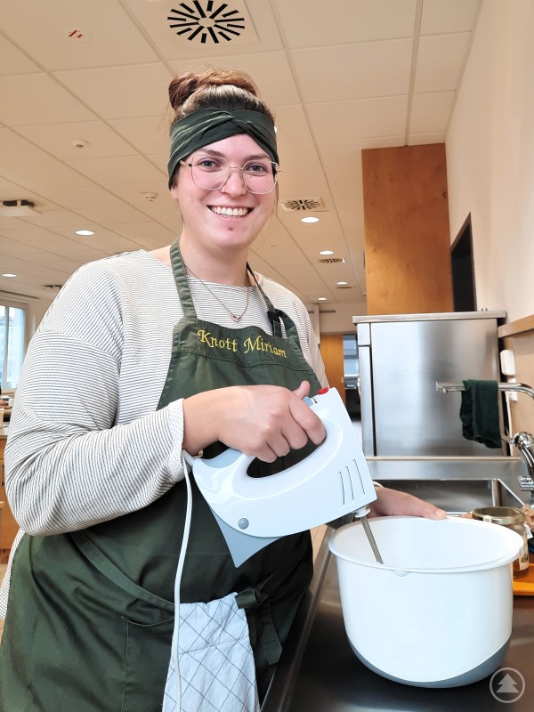 Die Herstellung ist einfach und geht sehr schnell. Man weiß, was drinnen ist und kann auch saisonale und regionale Aspekte berücksichtigen, findet Miriam Knott, Studierende der Fachschule in Passau. Eine junge Frau mit Schürze und Stirnband steht in einer Küche und rührt mit einem Handmixer Teig in einer Schüssel.