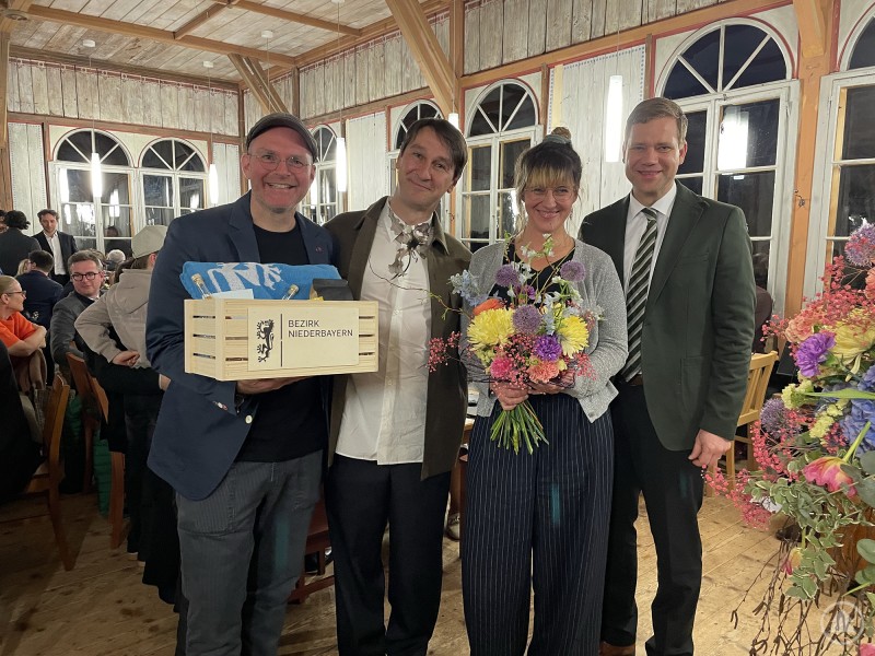 Bezirkstagspräsident Dr. Olaf Heinrich v.r. dankte Kuratorin Lisa Späthe, dem Präsidenten der Leir Foundation Markus Fuchs und Projektleiter Konrad Obermeier. Vier Personen stehen bei der Eröffnung einer Sonderausstellung im Freilichtmuseum Finsterau zusammen, eine Person hält Blumen, eine andere ein Geschenk des Bezirks Niederbayern in der Hand.