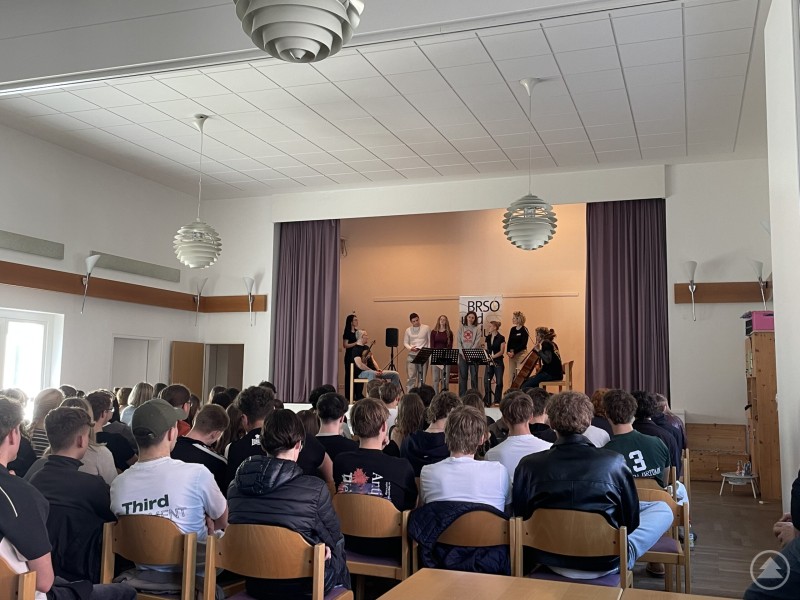 Musik und Hintergrundwissen verbinden sich zu einem eindrucksvollen Erlebnis für die Schülerinnen und Schüler. Schülerinnen und Schüler sitzen im Saal und verfolgen aufmerksam eine musikalische Darbietung auf der Bühne, bei der Musiker und Moderatoren im Einsatz sind.