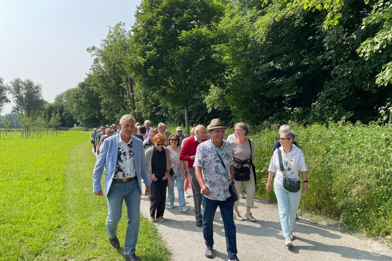 Bei einem Spaziergang durch die farbenfrohe Gartenwelt der Oö. Landesgartenschau kamen alle auf ihre Kosten. Eine Gruppe älterer Menschen spaziert bei Sonnenschein auf einem Weg entlang einer grünen Wiese und bewaldeten Kulisse.