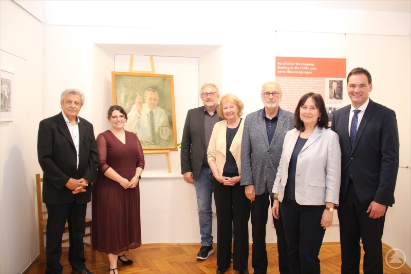 Gruppenfoto bei der Vernissage zur Franz-Schumertl-Ausstellung: Karl-Heinz Paulus, Gabriela Rauscher, Thomas Schumertl, Angelika Schumertl, Wolfgang Schumertl, Helma Schumertl und Landrat Sebastian Gruber (v. l.). Sieben Personen stehen in einer Ausstellung vor einem Porträt von Franz Schumertl, darunter Landrat Sebastian Gruber und Familienangehörige Schumertls.