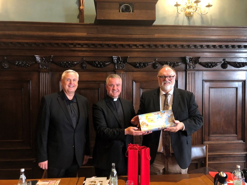 Oberbürgermeister Jürgen Dupper (rechts) zusammen mit Abt Patrick Schöder (Mitte) und Dompropst Dr. Michael Bär beim Empfang im Kleinen Rathaussaal. Drei Männer stehen nebeneinander in einem historischen Rathaussaal mit dunkler Holzvertäfelung; der mittlere Mann im Priestergewand hält mit dem Mann rechts ein Geschenkpaket.