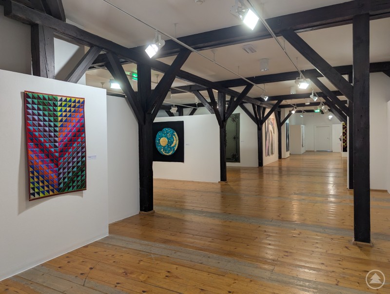 Farbenfrohe und vielfältige Quilt-Kunstwerke im Dachgeschoss des Stadtmuseums Deggendorf. Blick durch einen hellen Museumsflur mit schwarzen Holzträgern, an den Wänden hängen farbenfrohe, geometrische und abstrakte Quilts.