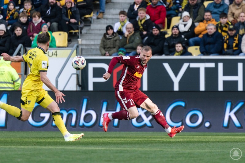 Ein Jahn-Spieler im weinroten Trikot nimmt den Ball volley, während ein Spieler von Alemannia Aachen im gelben Trikot versucht, den Abschluss zu verhindern. Im Hintergrund sind die Zuschauerränge gut besucht.