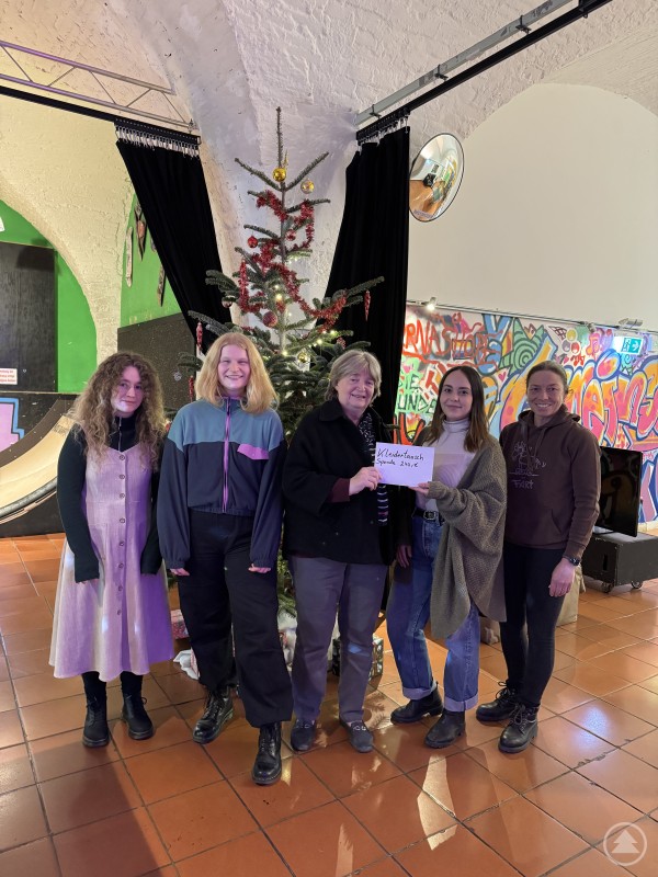 Beim Kleidertausch im Jugendzentrum Zeughaus konnten 240 Euro gesammelt und an das Frauenhaus Passau übergeben werden. Sechs Frauen stehen in einem Veranstaltungsraum vor einem geschmückten Weihnachtsbaum. Zwei von ihnen halten ein Schild mit der Aufschrift „Kleidertausch Spende 240 €“. Im Hintergrund sind farbenfrohe Graffiti-Wände und Vorhänge zu sehen, die Atmosphäre wirkt freundlich und gemeinschaftlich.