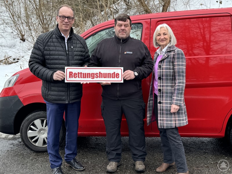 Vertreter der Rettungshundestaffeln der Region nehmen die Unterstützung entgegen und präsentieren das Schild Rettungshunde vor einem Einsatzfahrzeug. Drei Personen stehen vor einem roten Einsatzfahrzeug der Rettungshundestaffel und halten gemeinsam ein Schild mit der Aufschrift Rettungshunde.