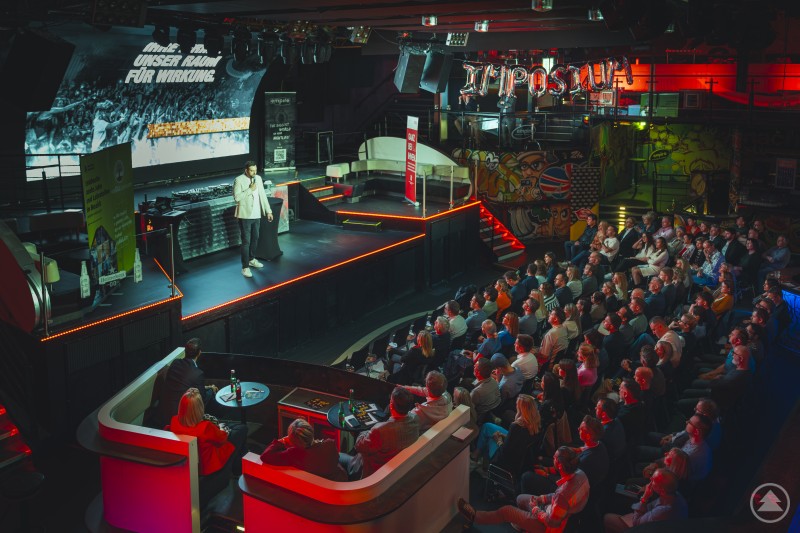 Gro&szlig;e Eventlocation mit B&uuml;hne und Publikum, ein Speaker steht unter Scheinwerfern und h&auml;lt einen Vortrag vor zahlreichen Zuh&ouml;rern.