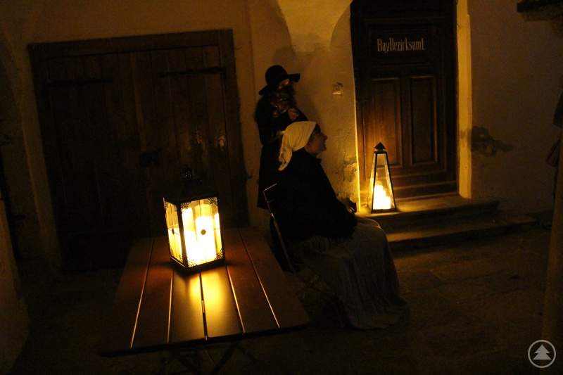 Unter den Arkaden vor der Türe des ehemaligen Bayerischen Bezirksamtes sorgte Laternenlicht für eine heimelige Stimmung. Judith Süß ist als Ehefrau des Wilderes im Vordergrund zu sehen, während die geheimnisvolle Gestalt (dargestellt von Gabriela Rauscher) im Hintergrund einen wachsamen Blick auf die Gäste hat.