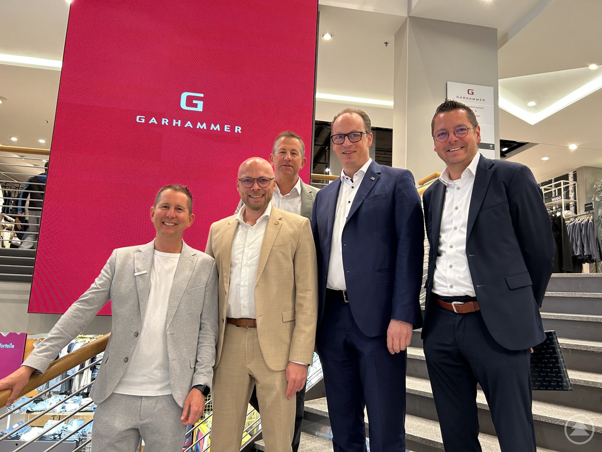 Digitalminister Fabian Mehring (2.v.l.) zeigte sich beeindruckt vom Modehaus GARHAMMER. Die Geschäftsführer Johannes (v.l.) und Christoph Huber führten auch MdL Martin Behringer und Bürgermeister Heinz Pollak über Laufstege und Showtreppen der textilen Begeisterung. Hier funktioniert vieles digital und verkauft sich echter Stoff dennoch hauptsächlich „analog“. Fünf Männer in Anzügen stehen vor einem großen pinkfarbenen GARHAMMER-Schild auf einer Treppe im Modehaus.