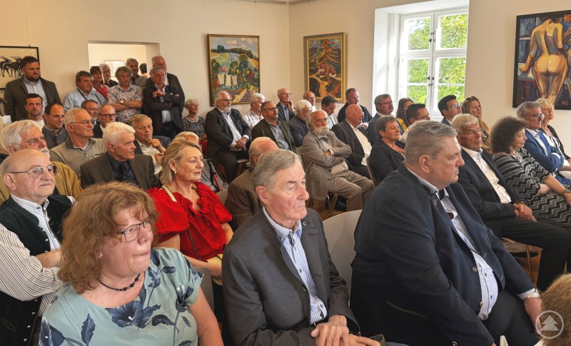 Zahlreiche Ehrengäste, Weggefährten und Bürger nahmen an der feierlichen Ausstellungseröffnung in der Galerie Wolfstein teil. Zahlreiche Gäste sitzen in einem gut besuchten Ausstellungsraum und folgen aufmerksam der Eröffnung. Im Hintergrund sind farbenfrohe Kunstwerke zu sehen.