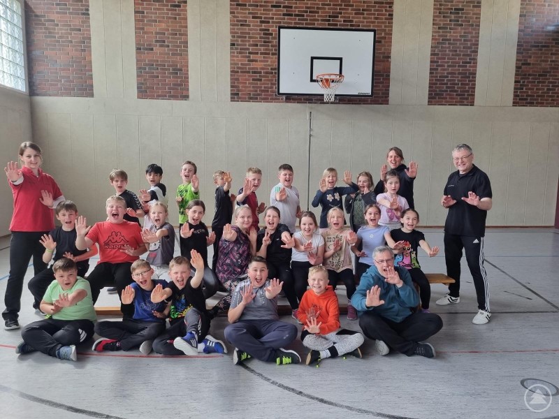 Eine große Gruppe von Kindern sitzt lachend in der Turnhalle und streckt demonstrativ die Hände nach vorne.