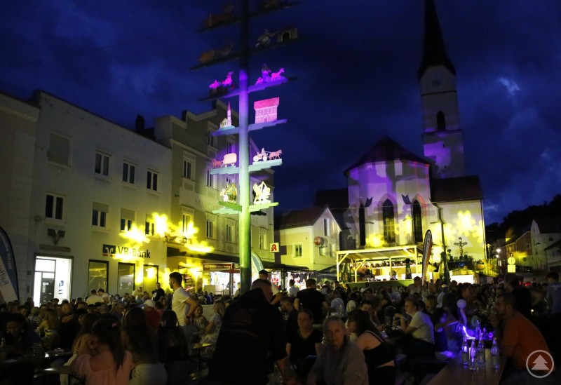 Magische Lichtshow und volles Haus beim Schönberger Marktfest: Der Marktplatz verwandelt sich in eine leuchtende Partymeile. Menschen sitzen dicht gedrängt an Biertischen am Abend; im Hintergrund eine Kirche mit bunter Lichtprojektion und ein dekorierter Maibaum.