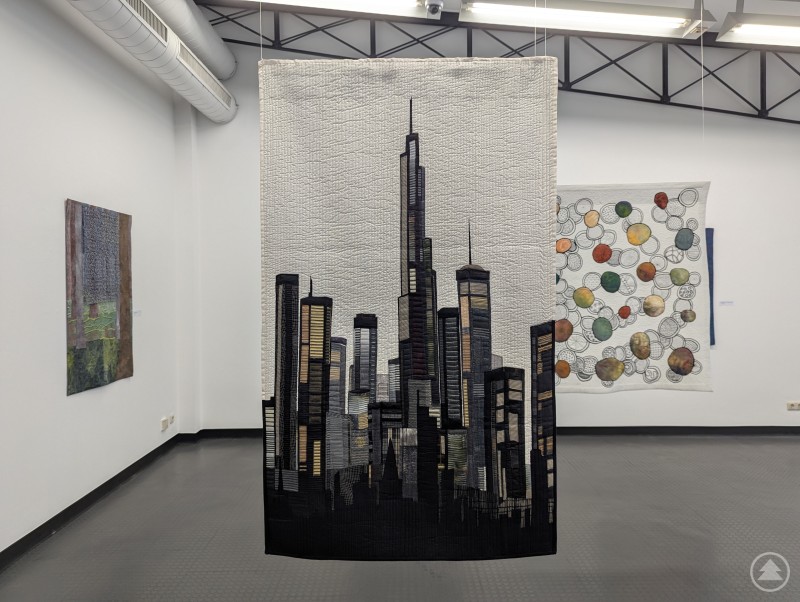 Ein moderner Quilt mit einer schwarzen Skyline vor hellem Hintergrund hängt frei im Raum einer Galerie, im Hintergrund sind weitere textile Kunstwerke zu sehen.