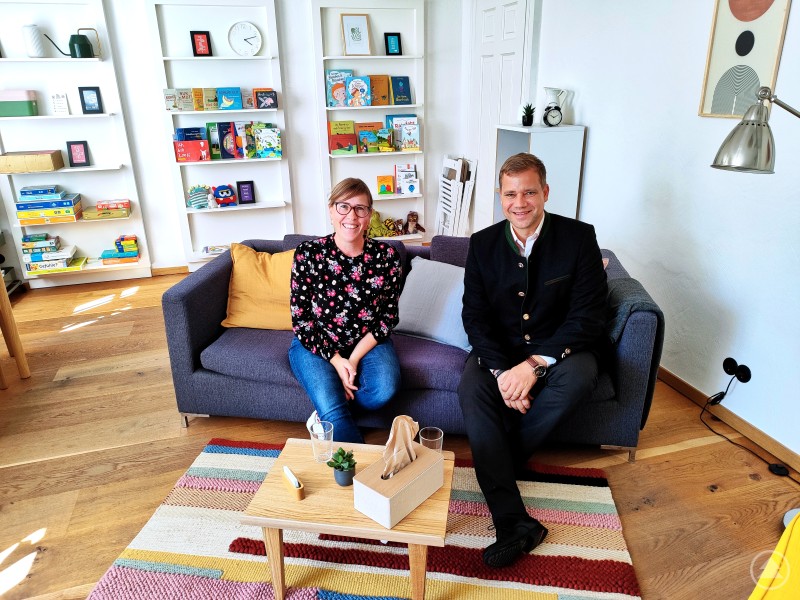 Nahmen auf der Couch der neuen Praxis für Kinder- und Jugendlichenpsychotherapie in Freyung Platz: Bezirkstagspräsident Dr. Olaf Heinrich (re.) und Therapeutin Maria Fleck (li.). Zwei Personen sitzen nebeneinander auf einem grauen Sofa in einer modern eingerichteten Praxis. Links eine Frau in Freizeitkleidung, rechts ein Mann im Anzug. Im Hintergrund sind weiße Regale mit Büchern und Spielen zu sehen.