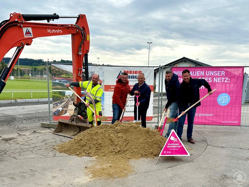 Eher symbolisch am Schaufeln: (v.li.) Vorarbeiter Manfred Mittendorfer, Geschäftsleiter Michael Braun, 1. Bürgermeister Robert Grabler, Bauleiter Matthias Heindl und Christian Lorenz von der Telekom. Fünf Männer stehen mit Schaufeln vor einem Sandhaufen neben einem orangefarbenen Bagger. Hinter ihnen sind Bauzäune mit Bannern der Telekom und der Baufirma Meier-Bau zu sehen. Die Stimmung wirkt positiv und symbolisiert den Start des Glasfaserausbaus.