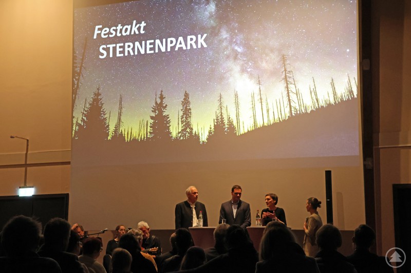 Naturpark-Geschäftsführer Hartwig Löfflmann (von links), Freyung-Grafenaus Landrat Sebastian Gruber und Nationalparkleiterin Ursula Schuster mit Moderatorin Julia Reihofer bei der Diskussionsrunde zur Sternenpark-Auszeichnung. Auf einer Bühne im Hans-Eisenmann-Haus diskutieren vier Personen vor einem projizierten Sternenhimmel mit der Aufschrift „Festakt Sternenpark“. Im Publikum sitzen zahlreiche Gäste, die dem Gespräch folgen.