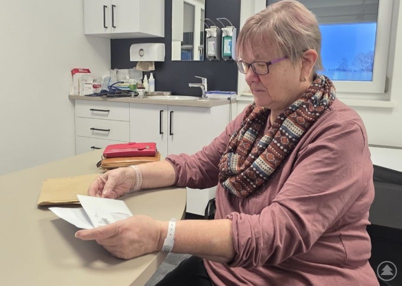 Manuela ist wieder zurück in der Heimat – die Erleichterung bei der Familie ist groß. Manuela sitzt in einem Behandlungsraum und liest Unterlagen. Sie trägt eine Brille und einen gemusterten Schal, im Hintergrund sind medizinische Utensilien zu sehen.