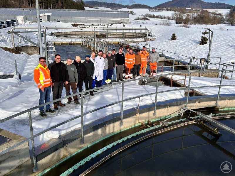 An zwei Tagen bereiteten sich angehende Fachkräfte für Abwassertechnik an der Kläranlage Schönberg praxisnah auf ihre Abschlussprüfung vor. „Team Orange“ aus dem Markt Schönberg erklärte dabei Abläufe, Technik und Anforderungen im Betrieb. Eine Gruppe von rund 15 Männern steht auf den Stegen einer Kläranlage im winterlichen Umfeld. Einige tragen orangefarbene Arbeitskleidung, andere Schutzkleidung oder Alltagsjacken. Im Hintergrund sind Becken der Abwasserreinigung, technische Anlagen sowie verschneite Wiesen und Hügel zu sehen.