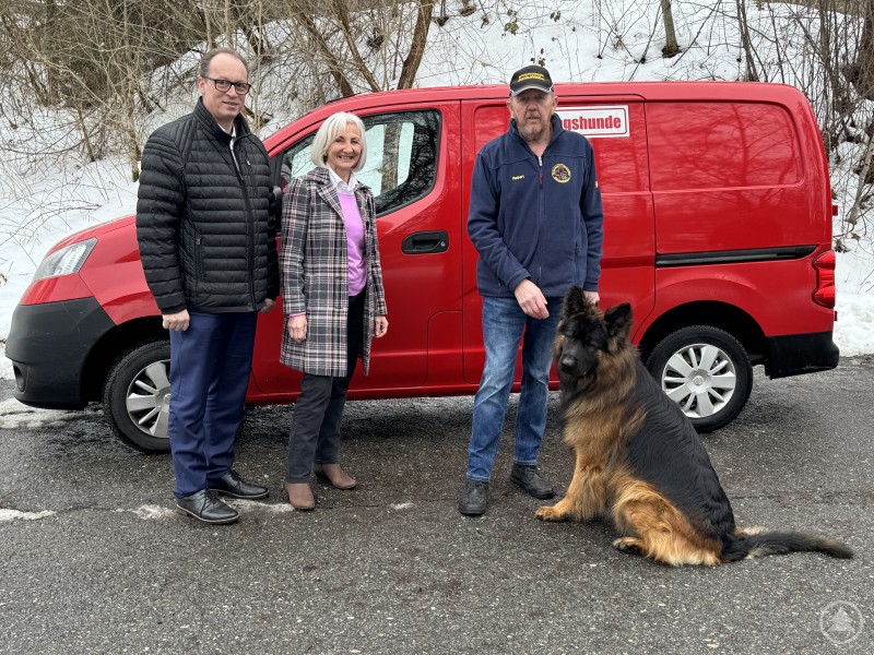 Mitglieder der Rettungshundestaffel präsentieren gemeinsam mit einem ausgebildeten Rettungshund ihre Arbeit vor einem Einsatzfahrzeug. Drei Personen und ein großer Deutscher Schäferhund stehen vor einem roten Fahrzeug der Rettungshundestaffel. Der Hund sitzt ruhig neben seinem Hundeführer.
