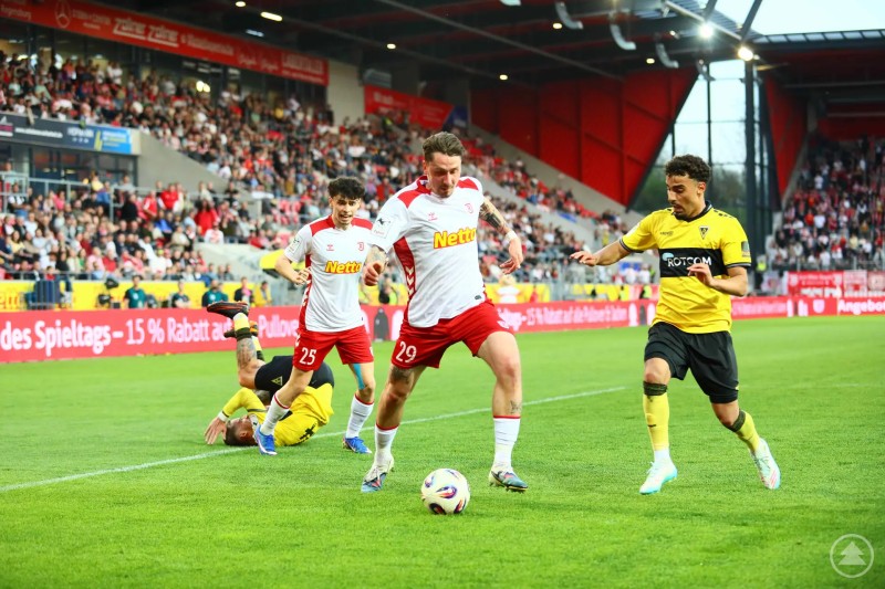 Eric Hottmann behauptet den Ball im Zweikampf und setzt sich energisch gegen mehrere Aachener durch. Zweikampf auf dem Spielfeld: Ein Spieler in Weiß-Rot führt den Ball, während ein Gegenspieler in Gelb attackiert. Ein weiterer Spieler liegt am Boden, das Stadion ist gut gefüllt.