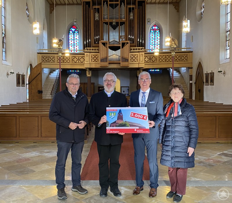 Landrat Josef Laumer, Pfarrer Alfons Leibl, Vorstandsvorsitzender Walter Strohmaier und Elfriede Kerscher bei der Übergabe des Spendenschecks in der Pfarrkirche Atting. Vier Personen stehen in einer Kirche vor einer großen Orgel und präsentieren gemeinsam einen symbolischen Spendenscheck. Im Hintergrund sind Kirchenbänke und farbige Fenster zu sehen.