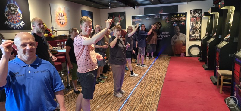 Mit viel Begeisterung und Teamgeist traten 56 Teilnehmer:innen bei der Dart-Meisterschaft der Caritas Werkstätten an. Eine Gruppe von Menschen mit und ohne Behinderung steht in einer Dart-Halle, bereit zum Wurf – sie lächeln und blicken motiviert in Richtung Zielscheibe.