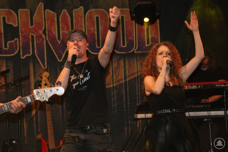 in Sänger und eine Sängerin der Band Rockwood performen leidenschaftlich auf der Bühne mit Mikrofonen, Bassgitarre und Keyboard im Hintergrund.