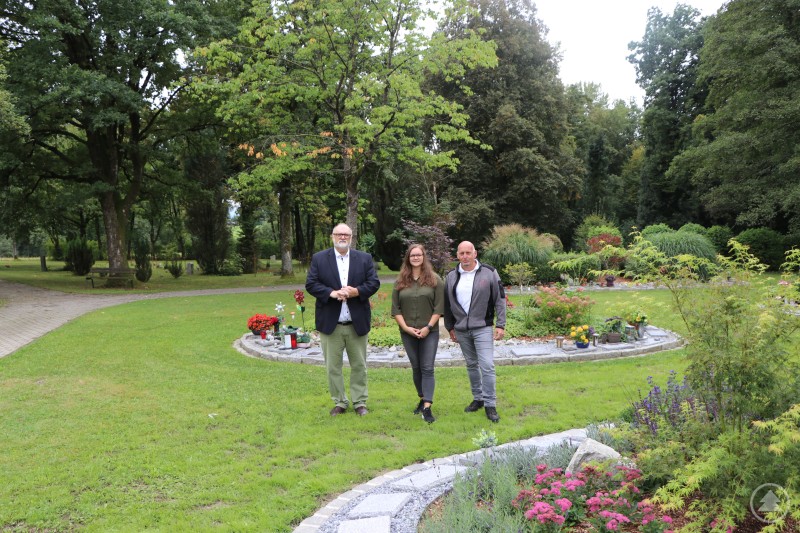 Oberbürgermeister Jürgen Dupper (links) besichtigt zusammen mit Stefanie Kocher von der Friedhofsverwaltung und Franz Urlberger, stellv. Leiter der Stadtgärtnerei, die neue Urneninsel am Friedhof St. Korona. Drei Personen stehen auf einer gepflegten Grünfläche des Friedhofs St. Korona, umgeben von bepflanzten Grabstellen und Bäumen.