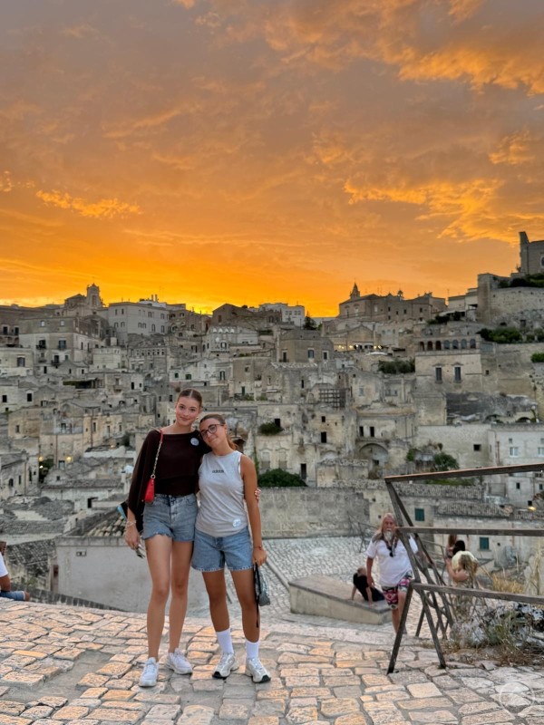 Hanna Lorenz (rechts) aus Schöllnach verbrachte im Rahmen des Rotary-Ferienaustauschs drei Wochen in Italien – eine unvergessliche Erfahrung. Zwei junge Frauen stehen in einer historischen Altstadt auf einer steinernen Terrasse. Hinter ihnen erstreckt sich eine beeindruckende Kulisse aus alten Gebäuden, während der Himmel in intensiven Orange- und Gelbtönen leuchtet. Beide lächeln und genießen den Moment bei Sonnenuntergang.