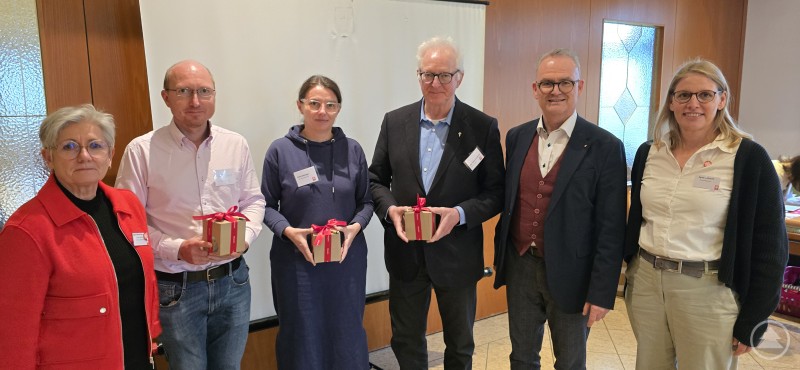 Neu in der Runde der Caritas-Vorsitzenden: Rita Bachinger (PCV Hunding-Lalling), Msgr. Kajetan Steinbeisser (St. Oswald), Pfarrer Matthias Grillhösl (Hinterschmiding) mit Bischöflich Beauftragtem Stefan Seiderer und Agnes Stefenelli von der Gemeindecaritas, sowie die Abteilungsleiterin Ingrid Aldozo (li.) Sechs Personen stehen in einem Raum nebeneinander und halten Geschenke mit roten Schleifen. Im Hintergrund ist eine Leinwand zu sehen.