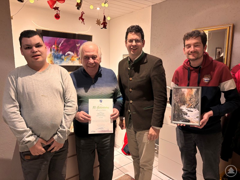 Pfarrer Simon Steinbauer und Bürgermeister Martin Pichler gratulierten Albert Preißer mit Bild und Glückwunsch zum 80er ebenso wie Enkel Manuel. Drei Generationen stehen zusammen in einem festlich geschmückten Wohnzimmer: Albert Preißer (Mitte, mit Urkunde), sein Enkel (rechts, mit Bild), Bürgermeister Martin Pichler (2. von rechts) und ein weiteres Familienmitglied. Die Stimmung ist herzlich und familiär.