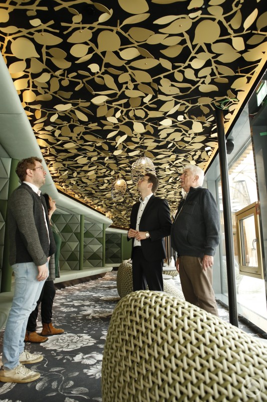 Unter dem gelaserten Blätterdach der neuen Oase der Stille im Familotel Schreinerhof: Jürgen (l.) und Günter (r.) Schon führten Veronika Egger und Bürgermeister Martin Pichler durch den neu gestalteten Ruhebereich. Vier Personen stehen in einem modern gestalteten Ruheraum mit auffälligem, goldfarbenem Blätterdach an der Decke. Die Gruppe blickt nach oben und betrachtet die architektonischen Details. Gepolsterte Sitz- und Liegebereiche sowie große Fenster sorgen für eine ruhige, elegante Atmosphäre.