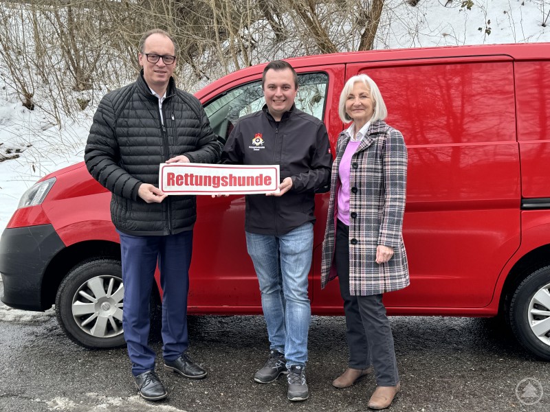 Vertreter der Rettungshundestaffeln zeigen das Rettungshunde Schild und freuen sich über die finanzielle Unterstützung für ihre ehrenamtliche Arbeit. Drei Personen stehen vor einem roten Fahrzeug der Rettungshundestaffel und halten ein Schild mit der Aufschrift Rettungshunde in die Kamera.