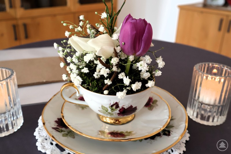 Frühlingsboten im antiken Kaffee-Service begrüßten die Gäste an den Tischen. Ein dekoratives Gesteck mit Tulpen und kleinen weißen Blüten in einer Tasse, daneben stehen Kerzen auf einem Tisch.