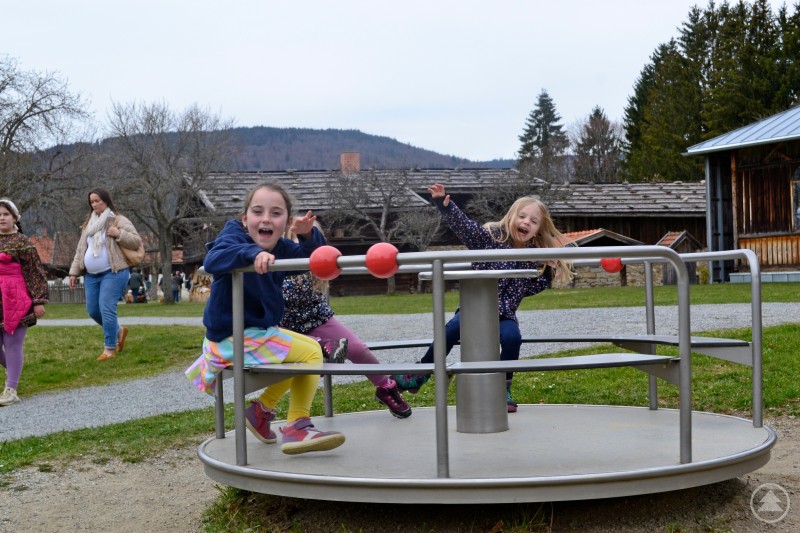 Spaß auf dem Spielpatz Das Bild zeigt fröhlich spielende Kinder auf einem Karussell im Freien – sie lachen, winken und genießen sichtbar den Moment. Im Hintergrund sind Erwachsene sowie weitere Kinder zu sehen, die über das Gelände schlendern. Die Umgebung mit den Holzhütten, der Naturkulisse und dem traditionellen Baustil deutet auf ein Freilichtmuseum oder einen ländlichen Veranstaltungsort hin. Die Szene vermittelt eine lebendige, familiäre Atmosphäre, passend zu einem Frühlingsfest oder Markt – vermutlich dem Eiermarkt 2025. Die Mischung aus Spiel, Natur und traditionellem Umfeld macht den Tag für Kinder wie Erwachsene zu einem Erlebnis.