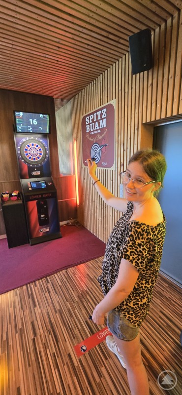 Eine Frau mit Brille und Leo-Shirt zielt konzentriert mit einem Dartpfeil auf eine elektronische Dartscheibe.