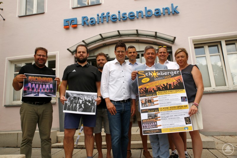 Das Organisationsteam mit Unterstützern der Raiffeisenbank bereitet das Schönberger Marktfest 2025 vor. Acht Personen stehen vor einer Raiffeisenbank, einige halten Plakate zum Marktfest Schönberg 2025 in der Hand.