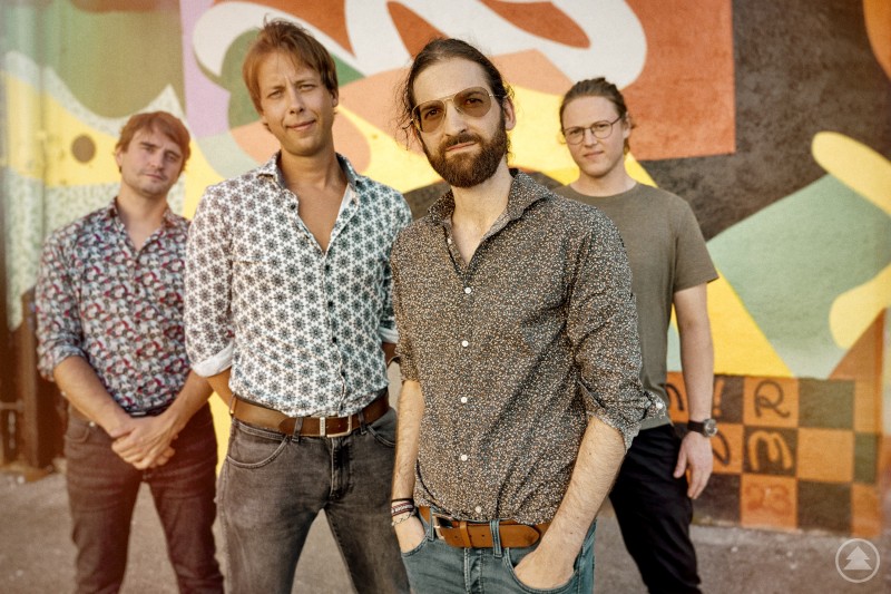 Joe Oldman Band - JOB1 Das Bild zeigt vier Männer, die als Band vor einer bunt bemalten Wand posieren. Sie tragen legere Hemden oder T-Shirts, zwei davon mit auffälligen Mustern. Einer der Männer steht leicht im Vordergrund, trägt Sonnenbrille und hat die Hände in den Hosentaschen.