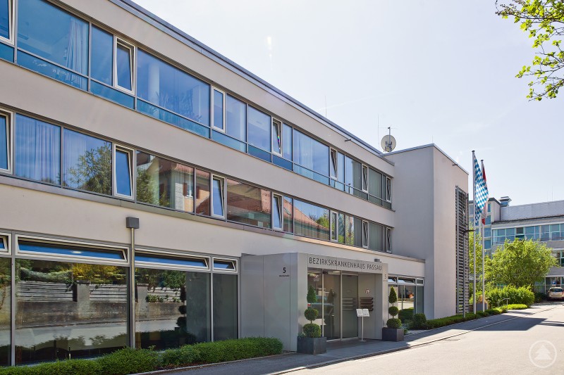 Das Bezirkskrankenhaus (BKH) Passau soll künftig enger mit der Kinderklinik Dritter Orden Passau im Bereich der Kinder- und Jugendpsychiatrie zusammenarbeiten. Das Foto zeigt die Außenansicht des Bezirkskrankenhauses (BKH) Passau. Das moderne, mehrstöckige Gebäude mit großen Glasfronten und hellem Putz steht an einer Straße, flankiert von Bäumen und einem gepflegten Eingangsbereich.